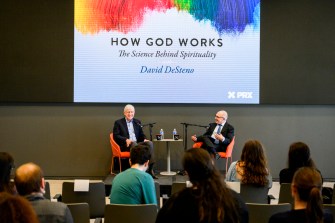 Dave DeSteno and Francis Collins speaking into microphones on stage in front of a screen that says 'How God Works' on it.