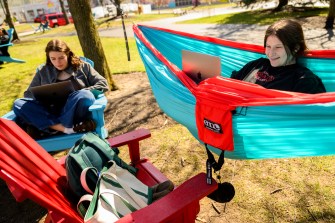 One student hanging in a blue and orange hammock while another sits in an adirondack chair, both using their laptops.