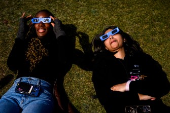 Two students laying on the grass looking up at the solar eclipse wearing eclipse glasses.