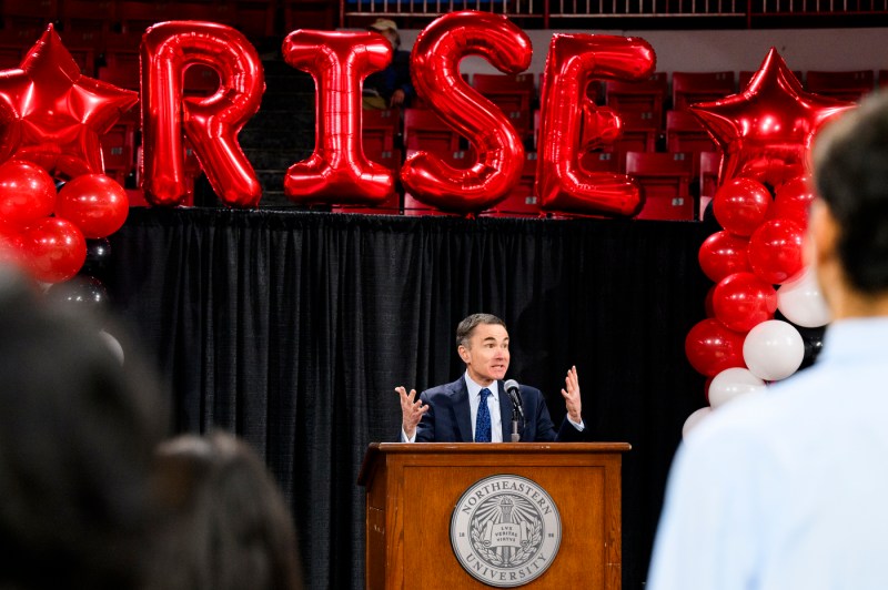 David Madigan speaking at a podium at the Rise EXPO under four red balloons that spell out the word 'RISE'. 