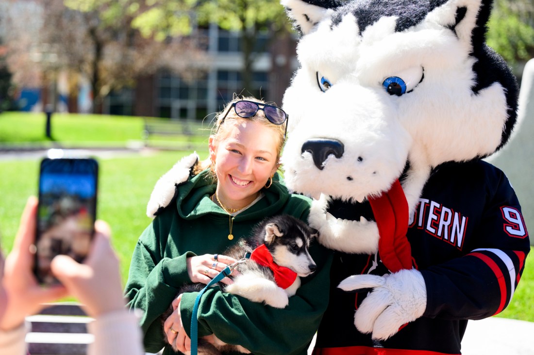 A person wearing a green sweatshirt cuddles a husky puppy while standing next to a husky mascot.
