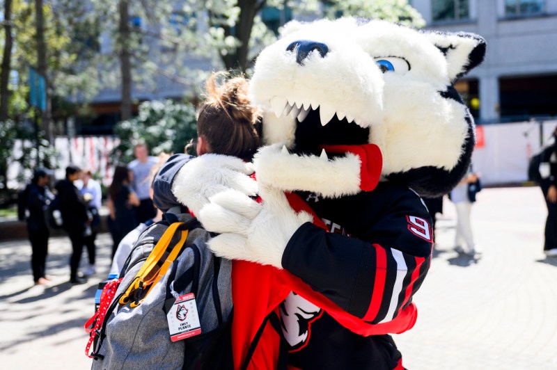 A husky mascot hugs someone carrying a grey-colored backpack outside on a sunny day.