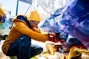 A person wearing a yellow jacket installs a purple-colored art piece on a wall.