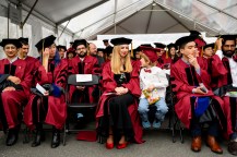 Graduates sit in rows of chairs under a tent dressed in regalia waiting to process.
