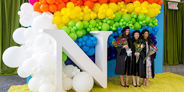 Graduates holding bouquets, standing under a rainbow arch of balloons next to a large letter N