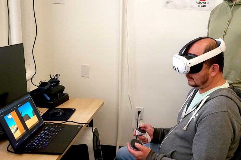 A man sitting in from of a laptop, wearing a VR headset and playing a game.