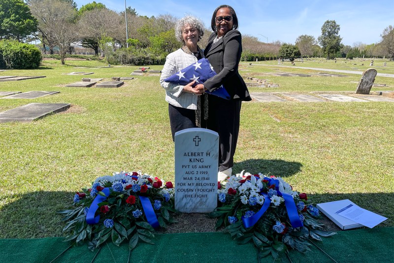 Two people smile and hold a folded American flag together as they stand behind Albert H King's grave. 