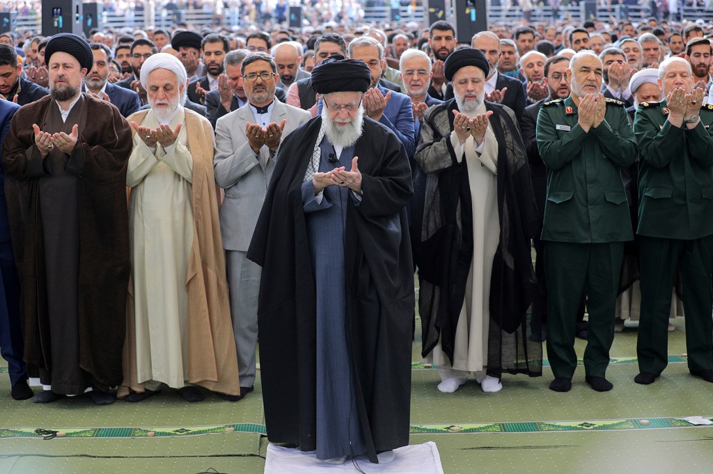 Ayatollah Ali Khamenei prays with raised hands in front of a large crowd, also praying.