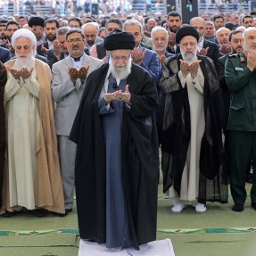 Ayatollah Ali Khamenei praying with hands uplifted in front of a large crowd, also praying.