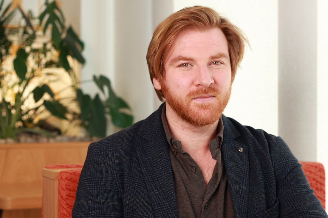 A man with red hair and beard wears a brown shirt and dark blazer. He sits in a red chair and a plant is over one shoulder.