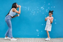 A teenage girl wearing jeans and a blue shirt blowing bubbles at a younger girl with pigtails.