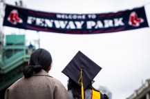A graduate wearing a cap and gown looking up at a 'Welcome to Fenway Park' banner outside of the park.