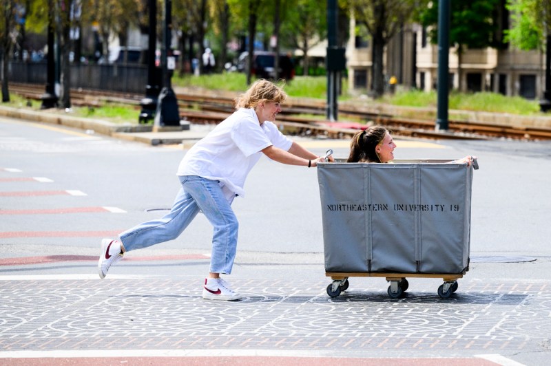 A person pushes another student in a moving bin across a street on a sunny day.