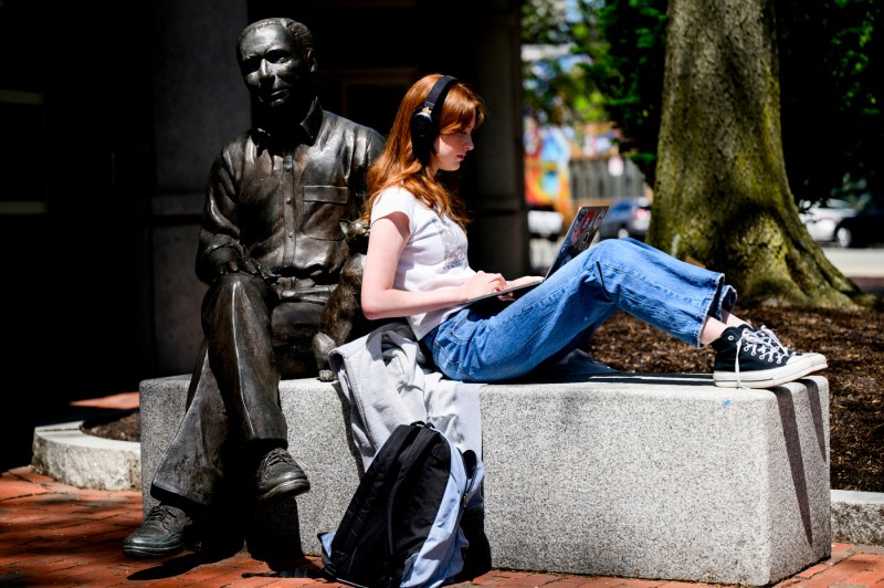A person works on their laptop while siting on a stone bench next to a statue on a sunny day.