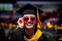 A graduate poses for a photo while placing one hand on the heart-shaped sunglasses that they are wearing.