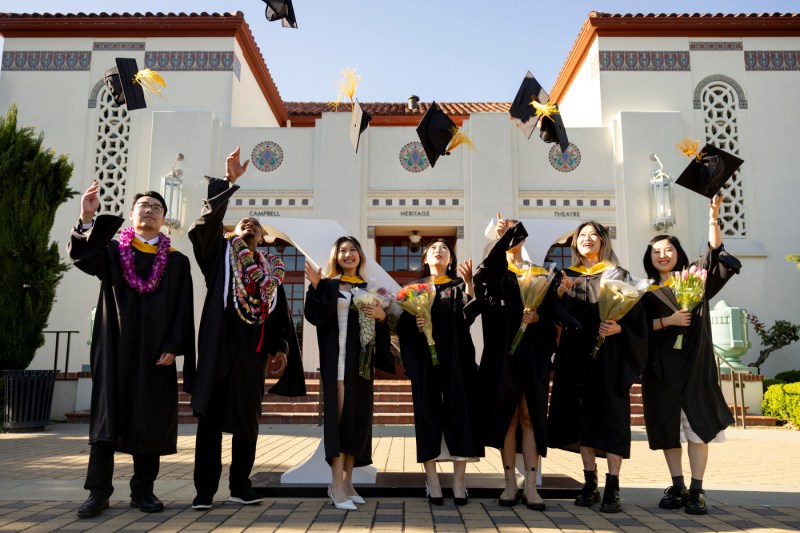 Seven people wearing regalia toss their mortarboards in the air while standing outside.