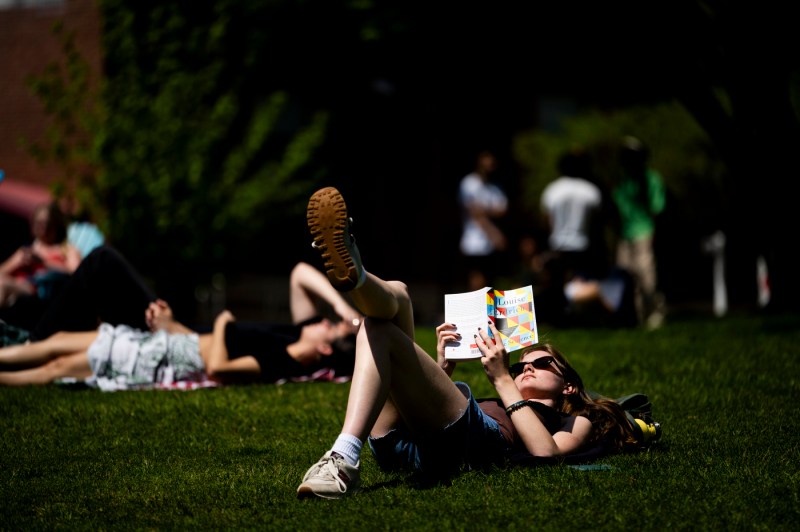 A person wearing sunglasses reads a book while lying in the grass outside on a sunny day.