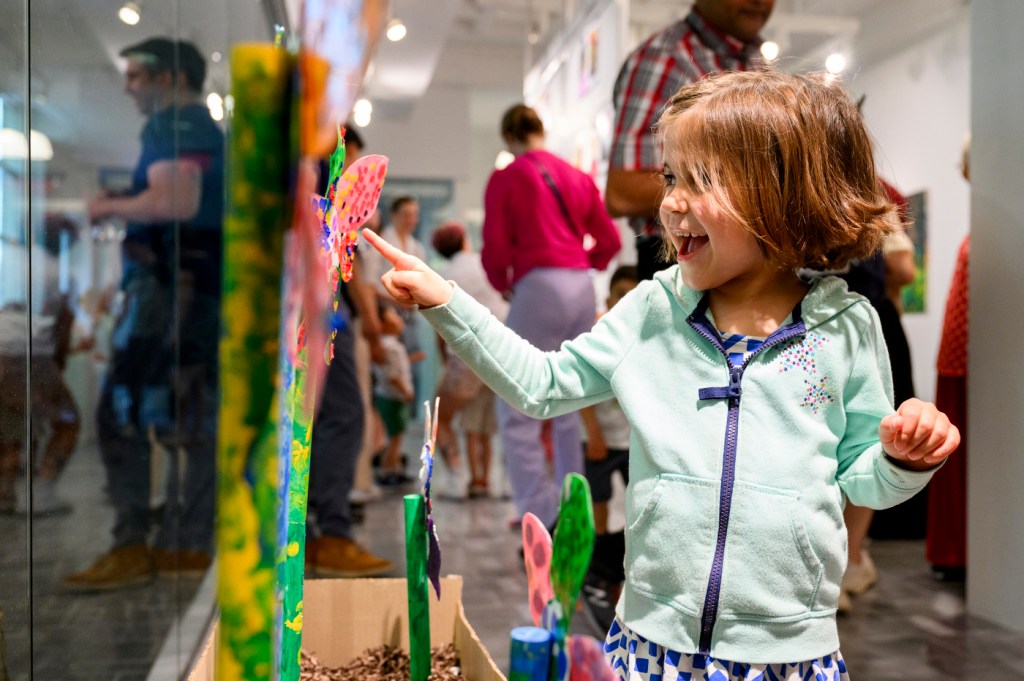 A child wearing a teal-colored sweatshirt points toward art on a wall.