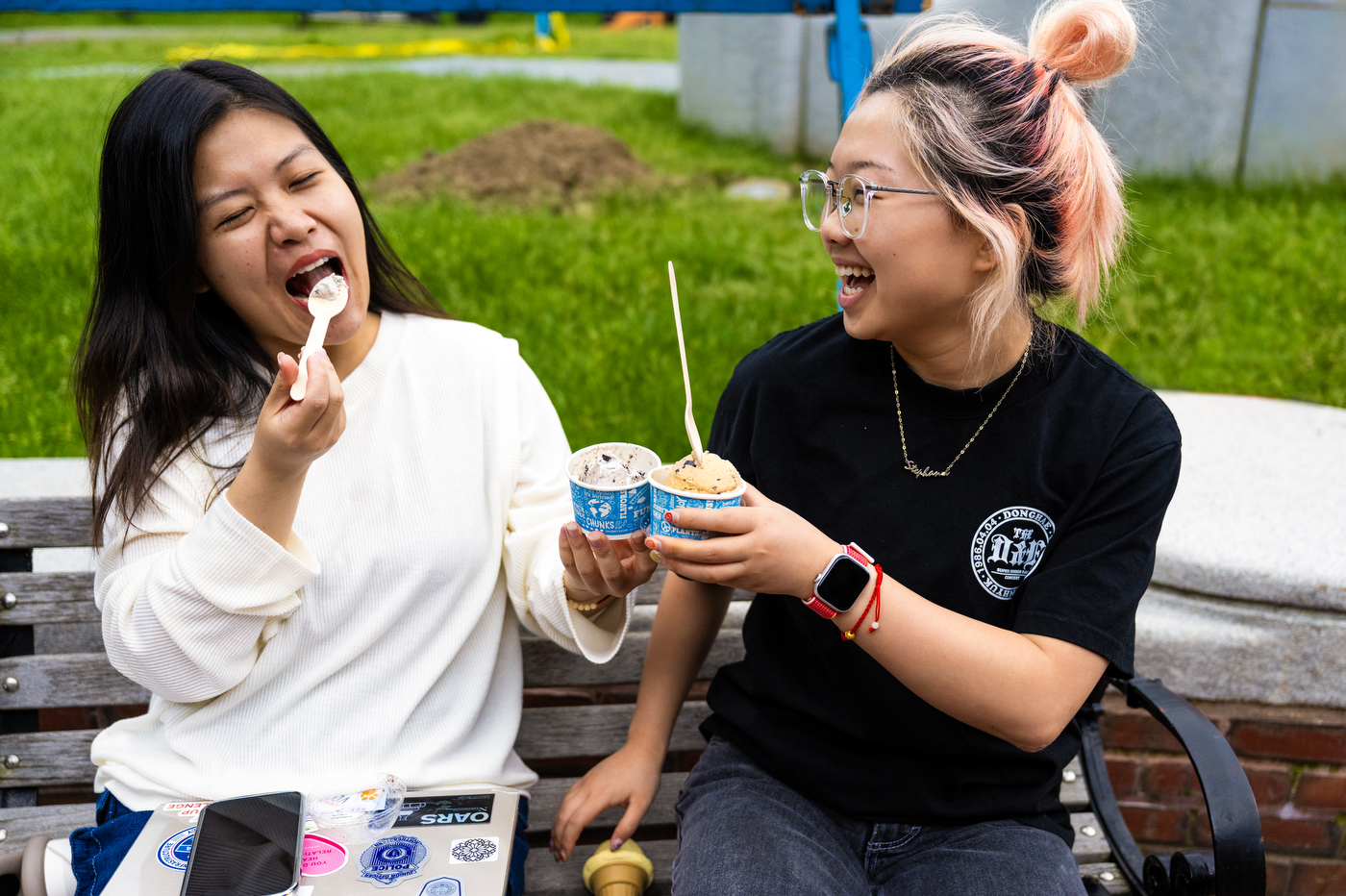 Two people laugh together while enjoying ice cream outside on a sunny day.