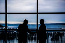 Silhouette of two people talking in front of floor to ceiling glass windows.