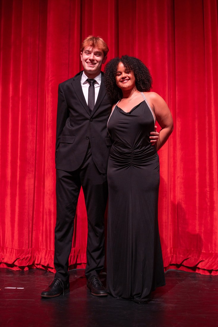 Two people posing in formal outfits in front of a red curtain.