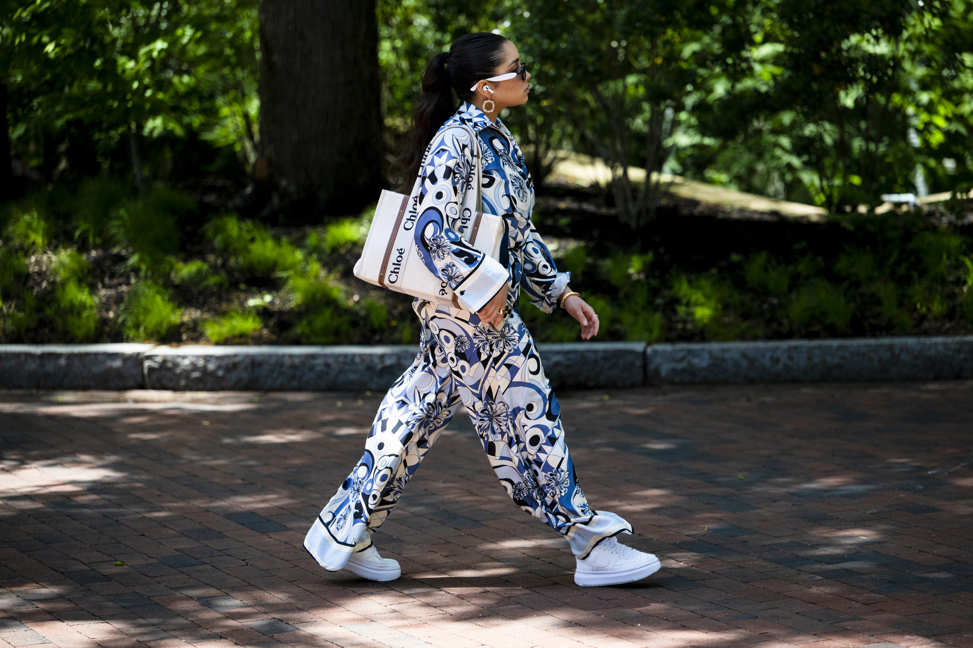 A person wearing a patterned white, blue, and black jumpsuit walks through a park on a sunny day.