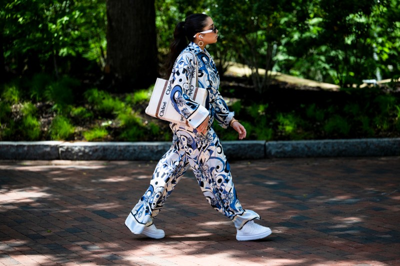 A person wearing a patterned white, blue, and black jumpsuit walks through a park on a sunny day.