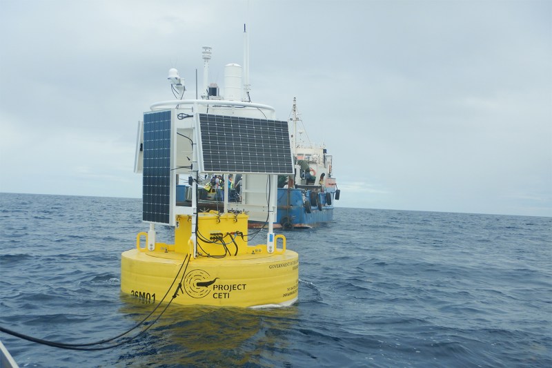 A yellow Project Ceti listening station that looks like a buoy with solar panels on it in the water near a boat.