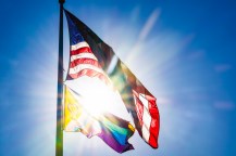 A LGBTQ Pride Flag flying underneath the US flag with the sun behind both.