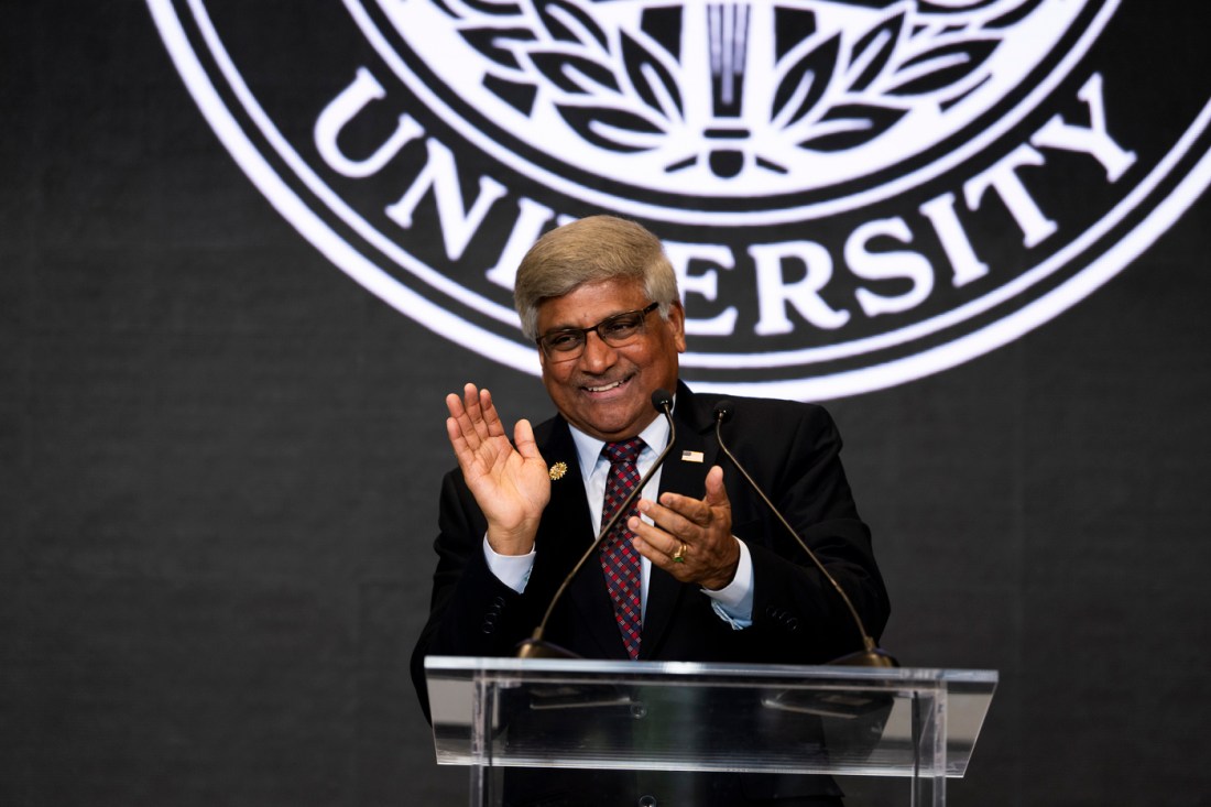 Sethuraman Panchanathan smiling and clapping at a podium, standing in front of the Northeastern University Seal.