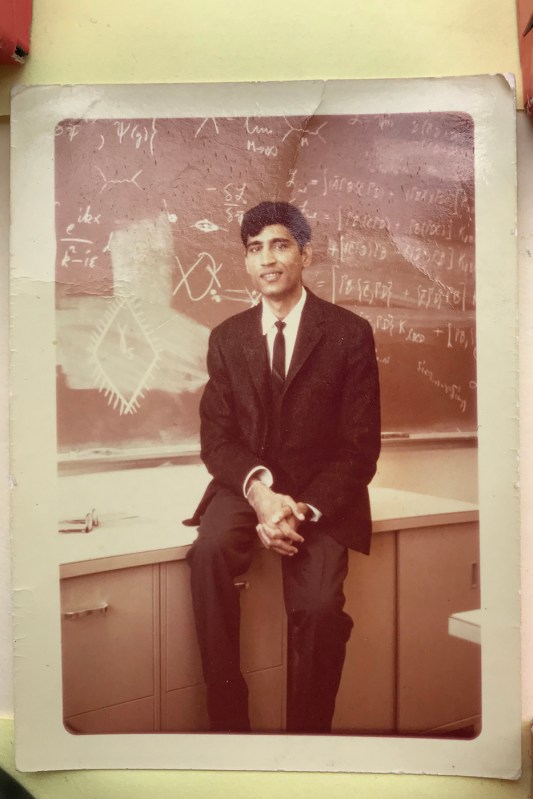 An old photo of a young Pran Nath sitting on top of a desk in front of a black board.