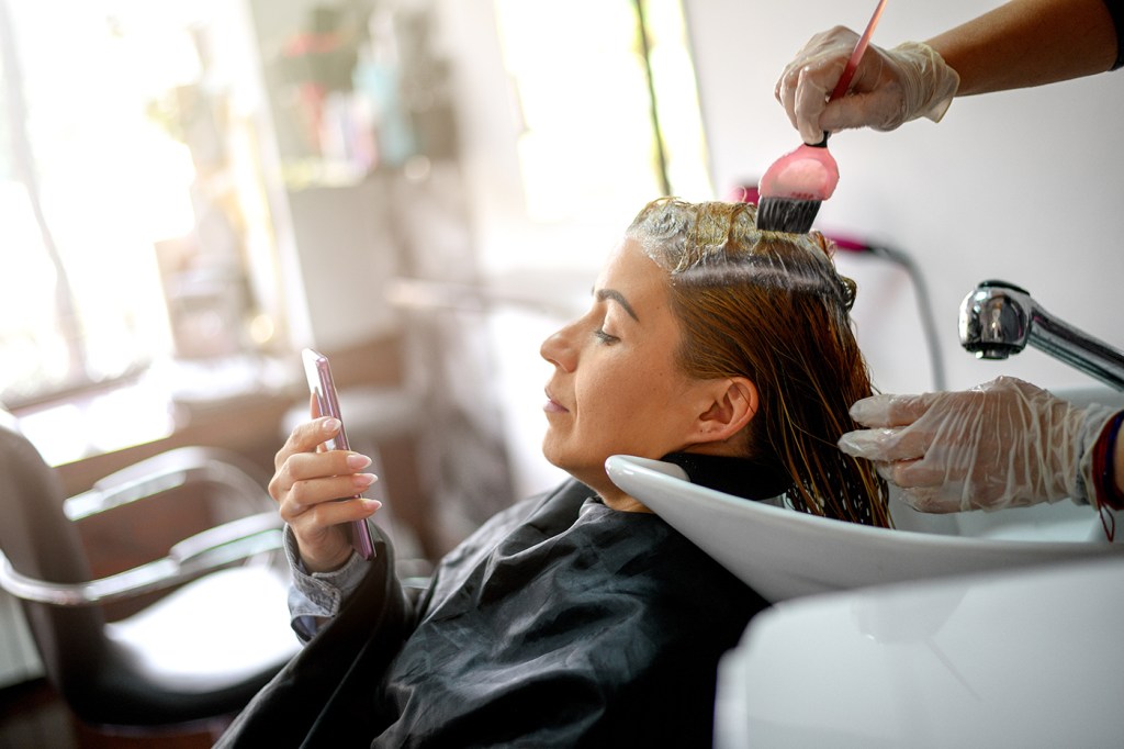 A woman getting her hair done at a salon while looking at her phone.