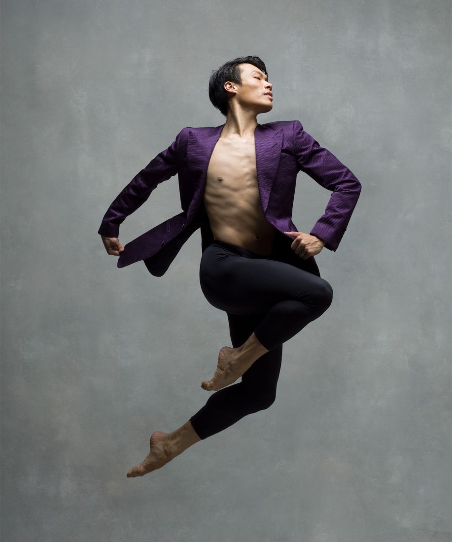 John Lam performing ballet, jumping midair wearing black pants and a purple blazer.