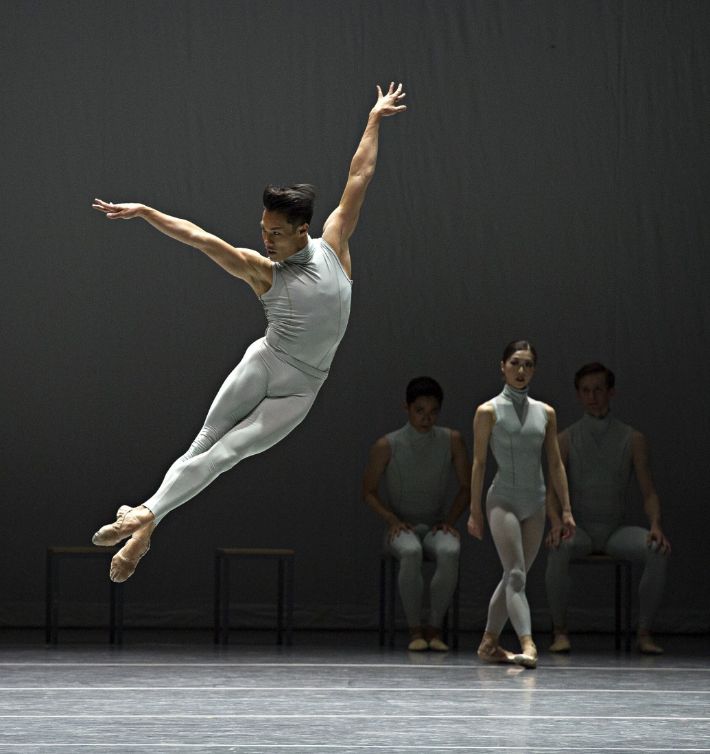 John Lam performing ballet, jumping in the air wearing a gray bodysuit. Other ballerinas wait in the background.