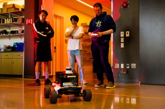 Three people stand around a four-wheeled robot in an orange-lit room.