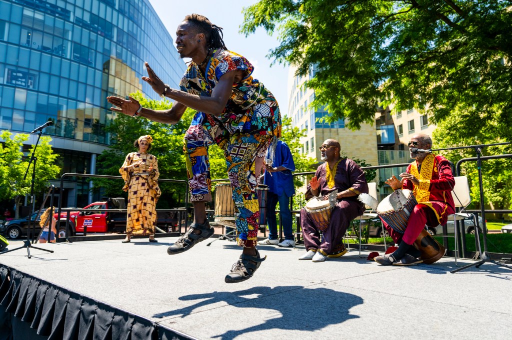 A dance and drum group performs on a stage outside on a sunny day.