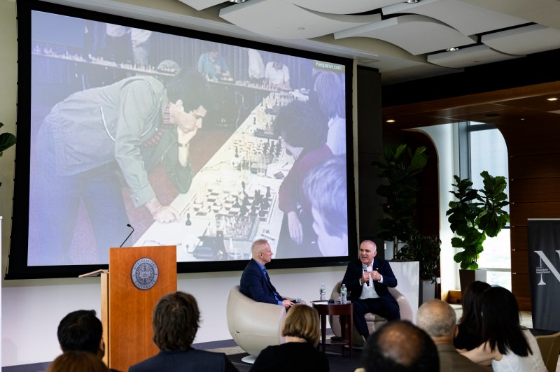 David De Cremer and Garry Kasparov sitting in chairs speaking in front of a PowerPOint slide showing a young Garry Kasparov playing chess.