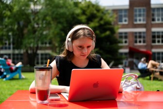 A person works on a laptop at a red table outside on a sunny day.