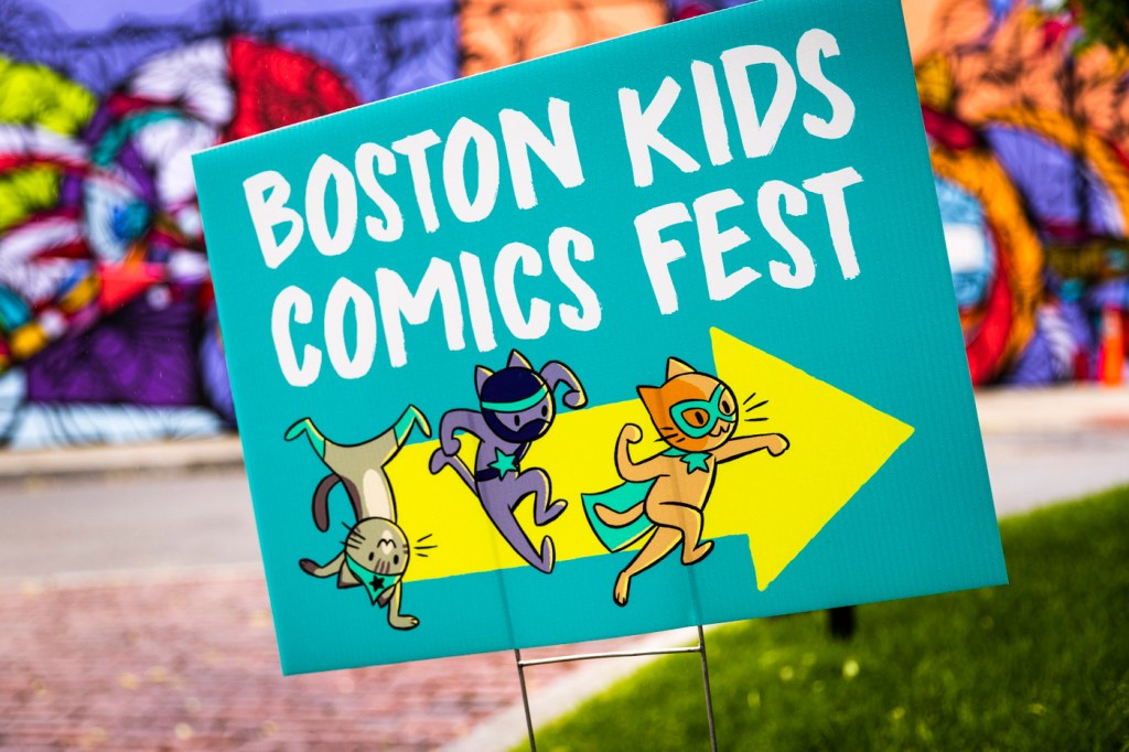 A turquoise sign that says 'Boston Kids Comics Fest' with a yellow arrow and three comic cats pointing to the right.