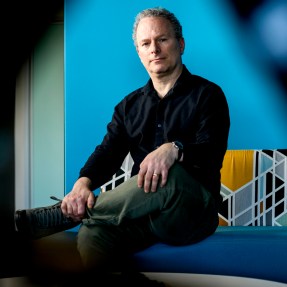 David Lazer sitting in a chair in front of a blue wall with one leg crossed over the other.