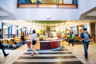 People walk around a brightly lit dining hall while holding trays.