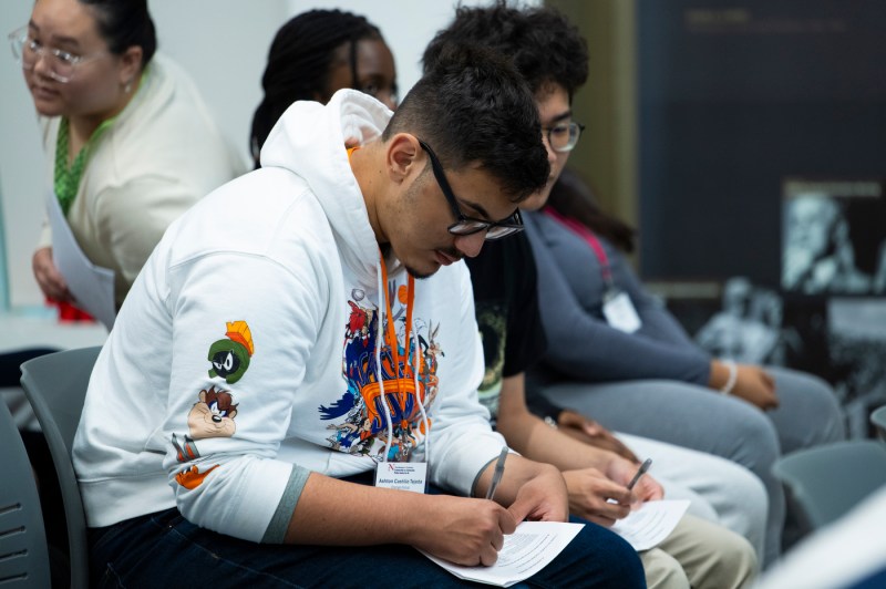 Close-up of a student taking notes with a pen and paper at the Northeastern Summer Youth Jobs Program orientation.