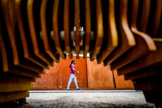 Seen through a wooden, semi-spherical, ridged cutout, a person walks across a sidewalk.