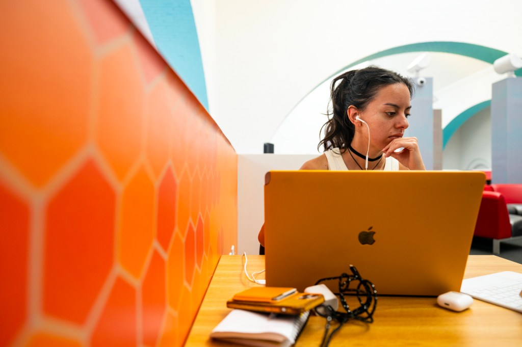 A person sits at a table with earbuds while working on a laptop.