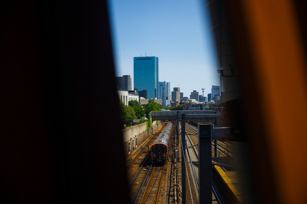 A train moves along its tracks in front of a city skyline, while being partially obscured on either side by shapes in the foreground.