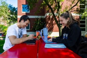 Two people work on their laptops while sitting at a bright red table outside on a sunny day.