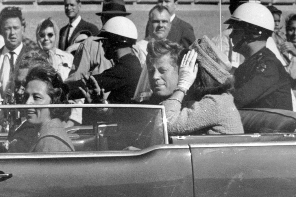 A black and white archival photo of President John F. Kennedy waving from his car in a motorcade.