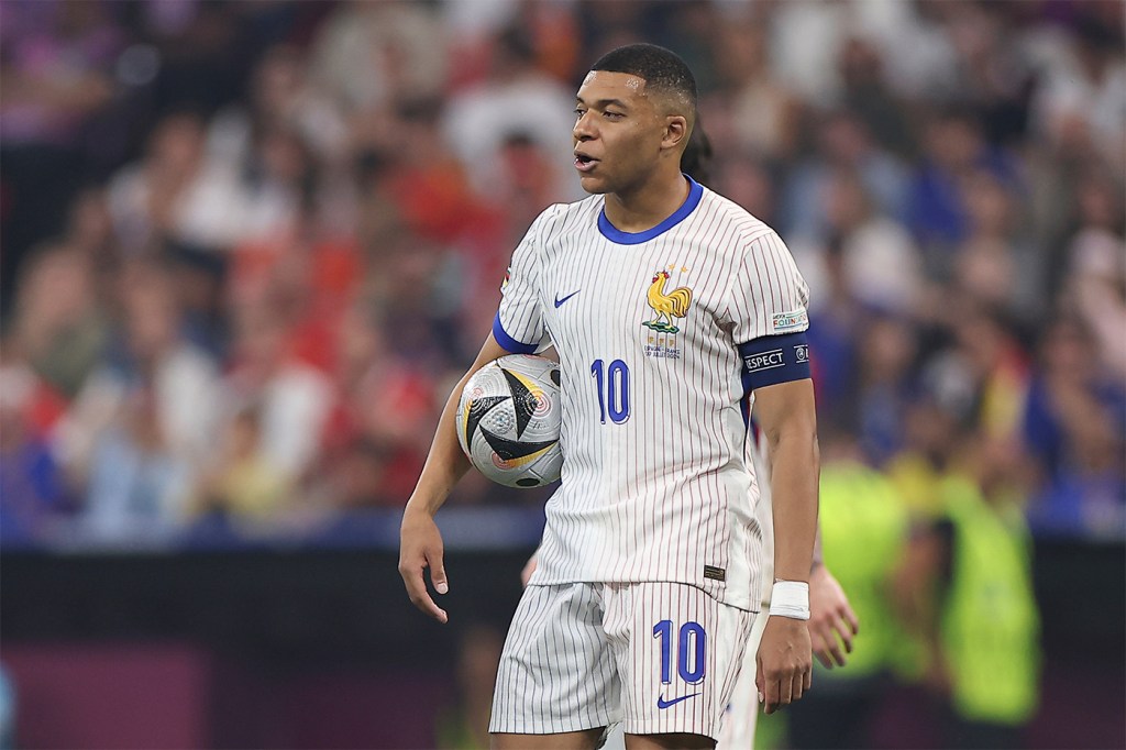 Kylian Mbappe in a white uniform holds a football under his right arm during a EURO 2024 match.