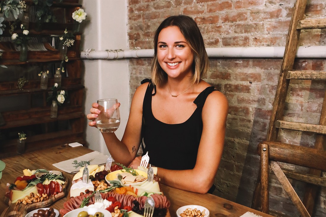 Marissa Mullen holding a glass of wine and smiling in front of one of her beautiful cheeseboards.