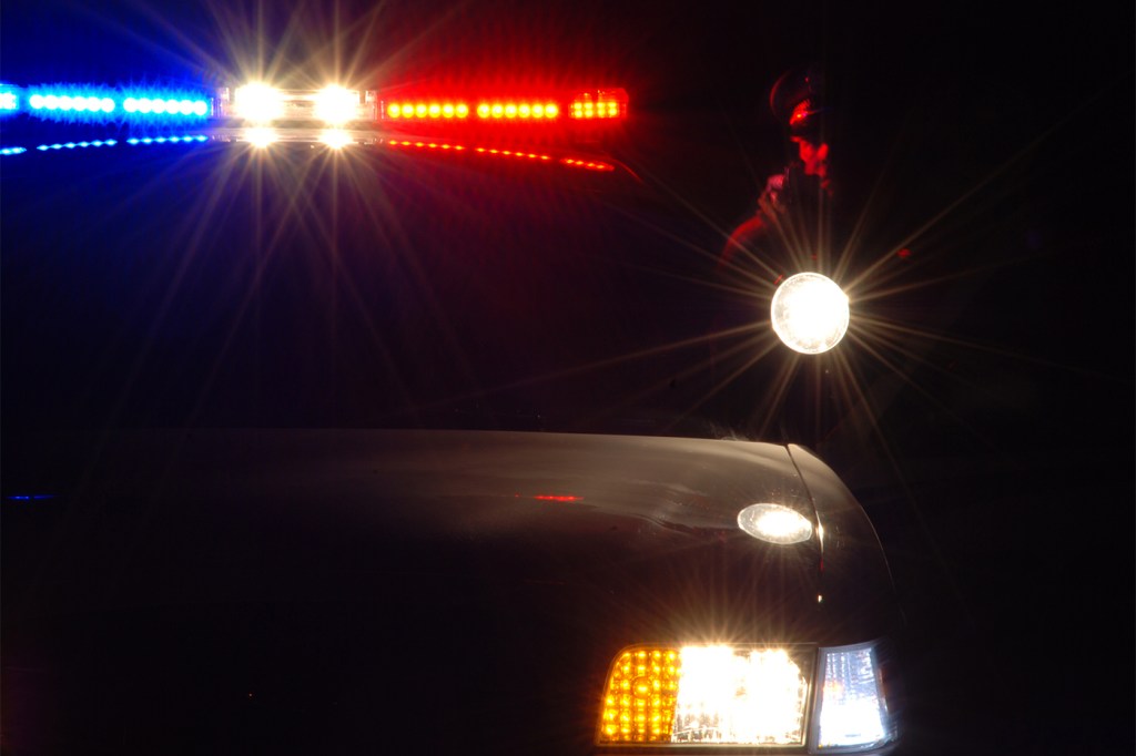 A police car's lights shine in the dark and illuminate the outline of an officer holding a flashlight.